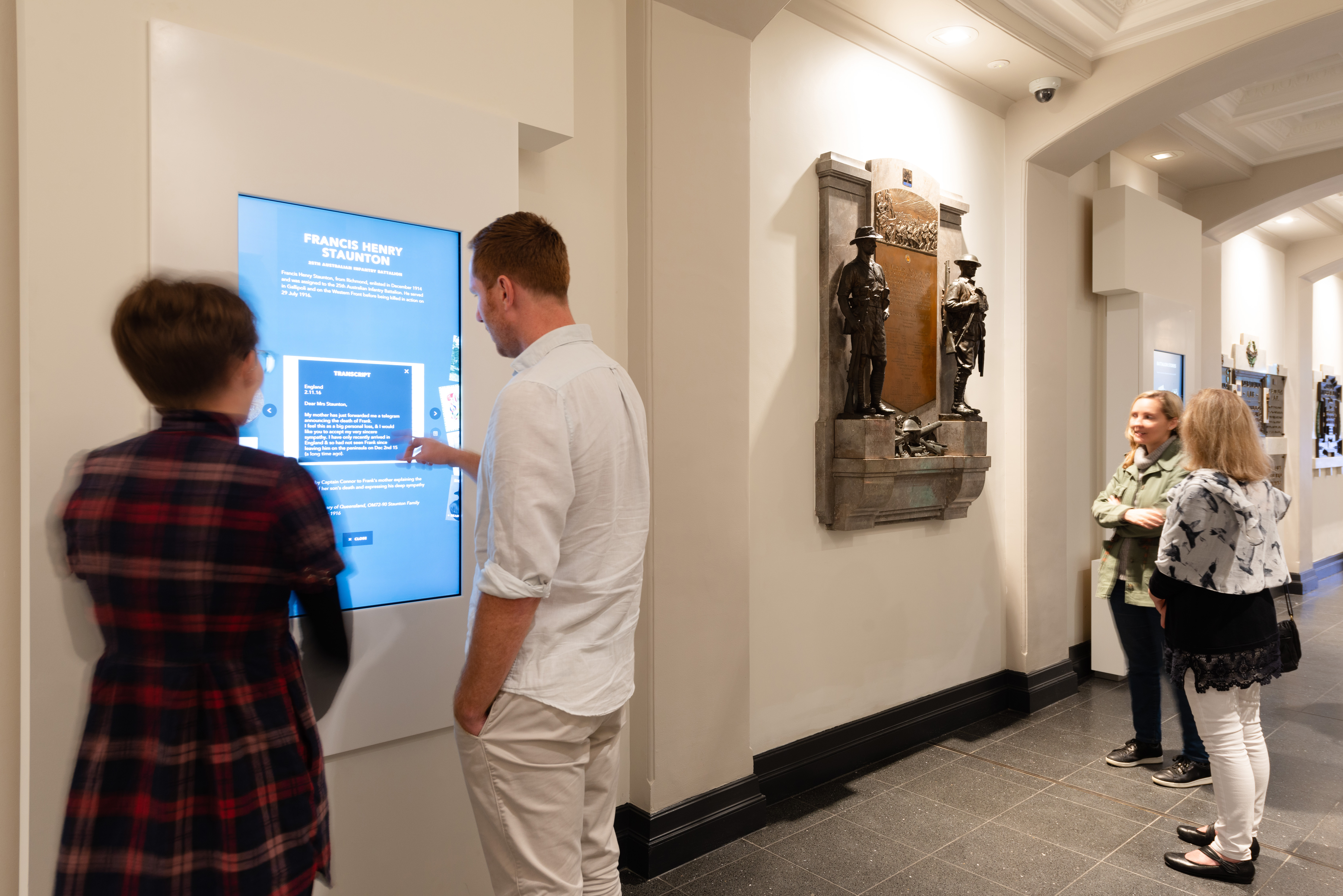 Two groups of people using a touch screen and viewing a memorial plaque.