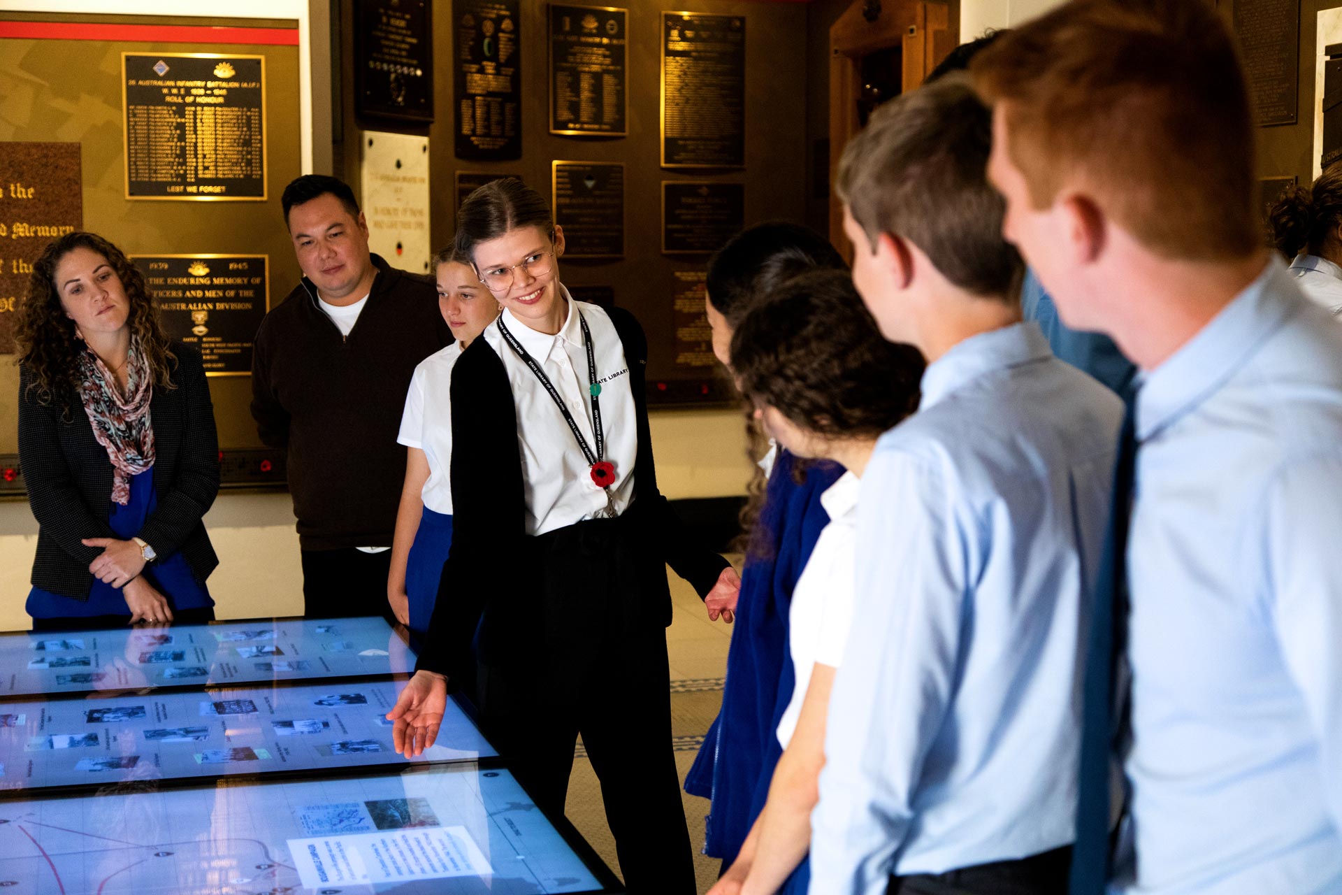 State Library staff member gesturing towards touch screens with a group of high school students. 