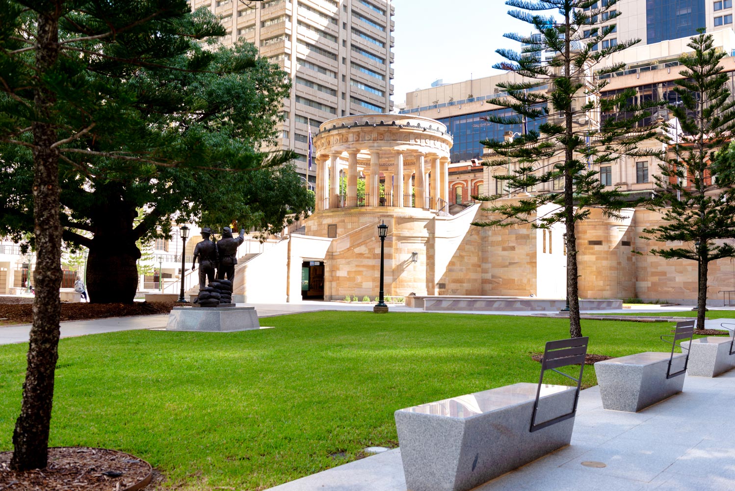 Anzac Square parkland with trees, a statue, benches, grass and the Shrine of Remembrance in the centre. 