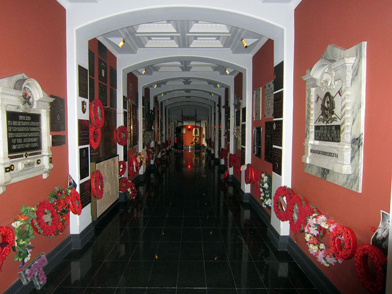 Colour image of interior corridor. Walls dark peach, smooth black tiles on floor, plaques and wreaths along wall.