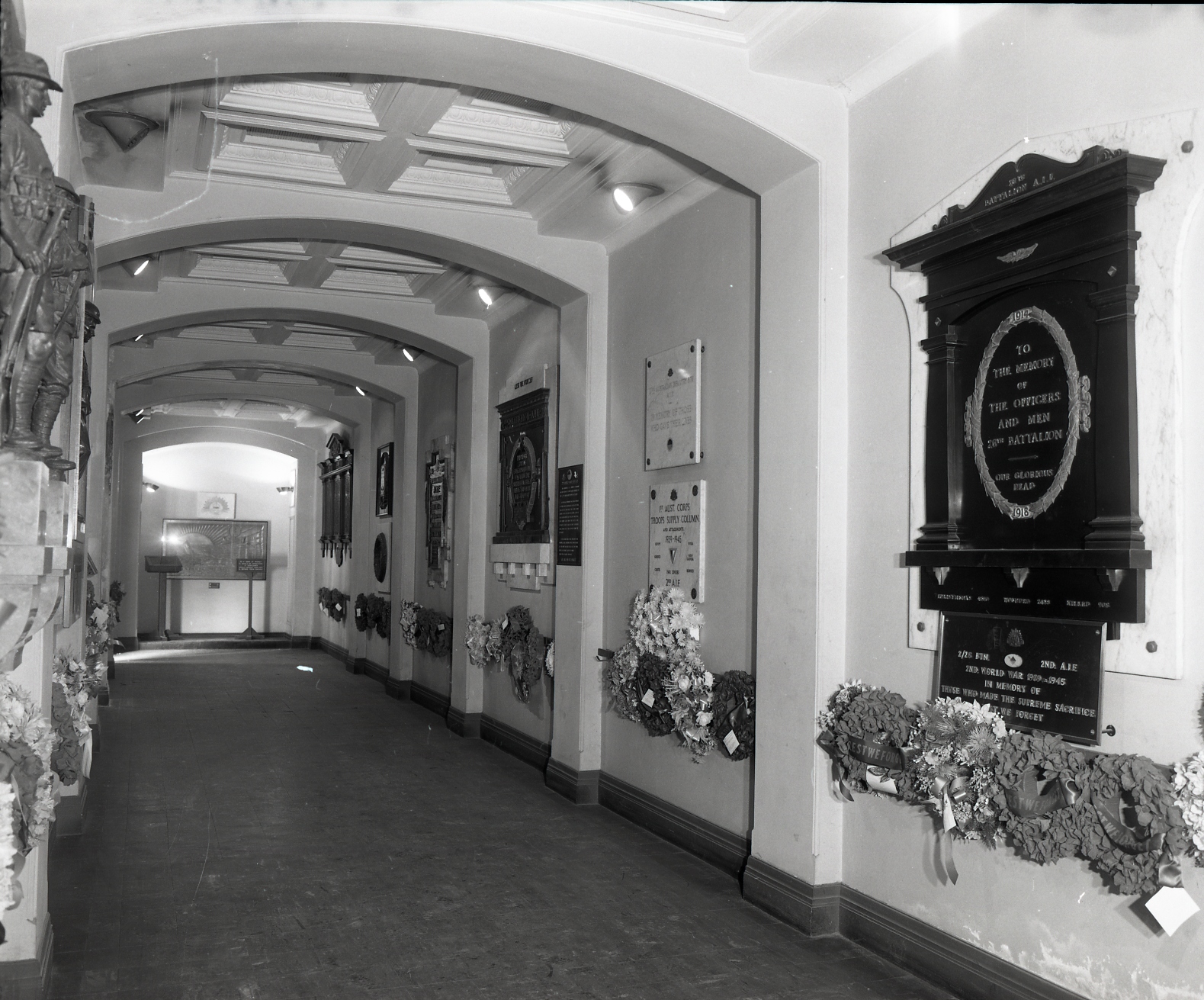 Interior black and white image of the crypt corridor focusing on the right hand side wall, showing wreaths and plaques.