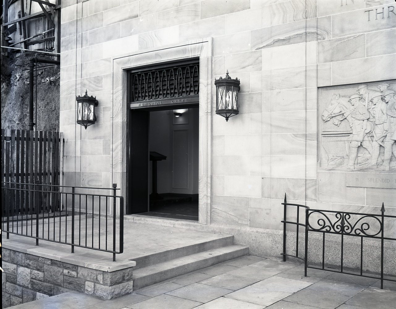 Black and white image taken from outside of the entrance to Anzac Memorial Crypt, showing two steps leading to the doorway and two lights attached to the wall either side of the entrance.