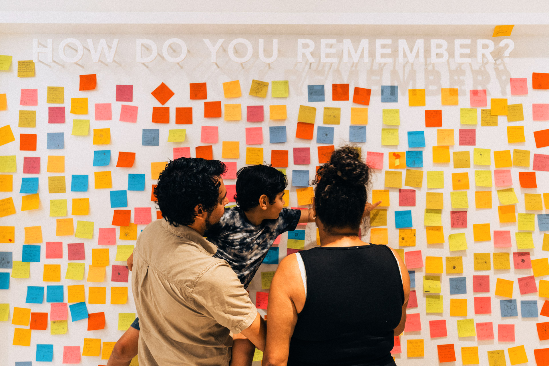 Woman, man and child standing in front of a wall covered in post-it notes. At the top of the wall large letters read 'how do you remember?'