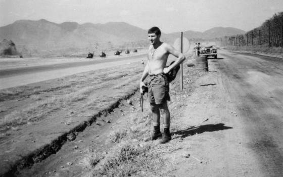 Corporal Victor Paul Patty, clad only in his shorts and boots, is standing alongside a road at Luscombe Field. Five helicopters are in a line on the 'landing field' to the side of him and a jeep with two men stood alongside it behind him.