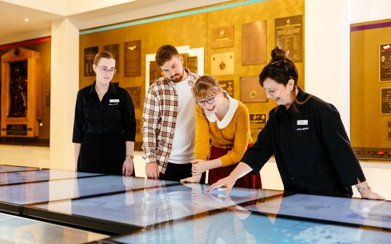 Two visitors use the interactive screens in the gallery with two staff members. 
