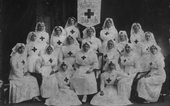 Old photograph of a group women nurses. Their uniforms feature a cross on the front and on the headdress. A sign in the middle reads 'Hamilton Branch.'