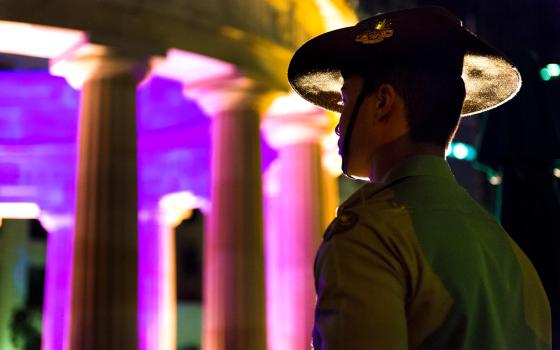 Young soldier wearing a slouch hat and standing at attention as he looks towards the Shrine of Remembrance at Anzac Square. The shrine is lit in pink and purple lights.