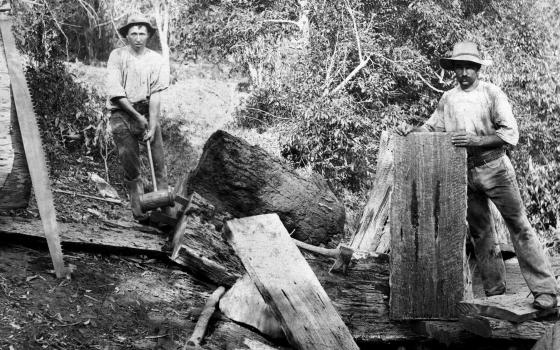 Black and white photograph of two men cutting large slabs of wood outside.
