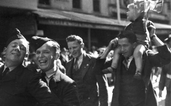 Black and while photograph of soldiers smiling as they walk down the street.  One of the men is carrying his child on his shoulders. The child is holding some Australian flags.