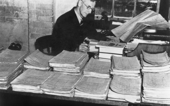 Black and white photograph of a man surrounded by papers on a desk.