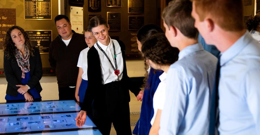 State Library staff member gesturing towards touch screens with a group of high school students.