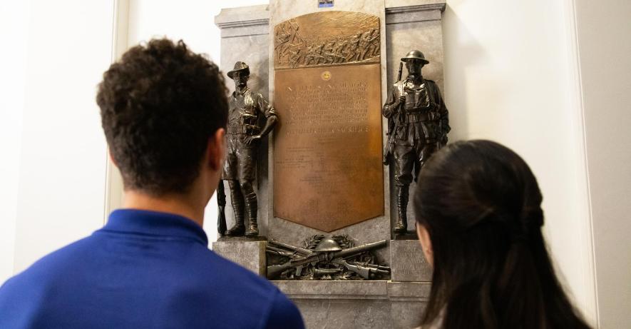 The back of two figures looking at a plaque featuring two soldiers in the gallery.