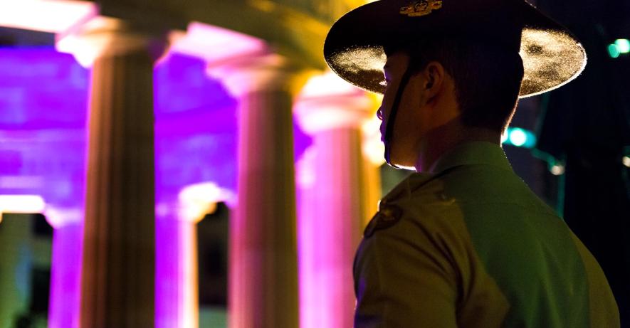 Young soldier wearing a slouch hat and standing at attention as he looks towards the Shrine of Remembrance at Anzac Square. The shrine is lit in pink and purple lights.