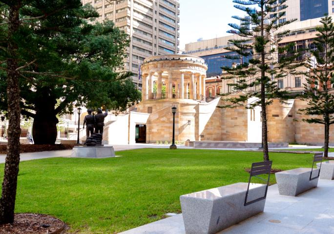 Anzac Square parkland with trees, a statue, benches, grass and the Shrine of Remembrance in the centre.