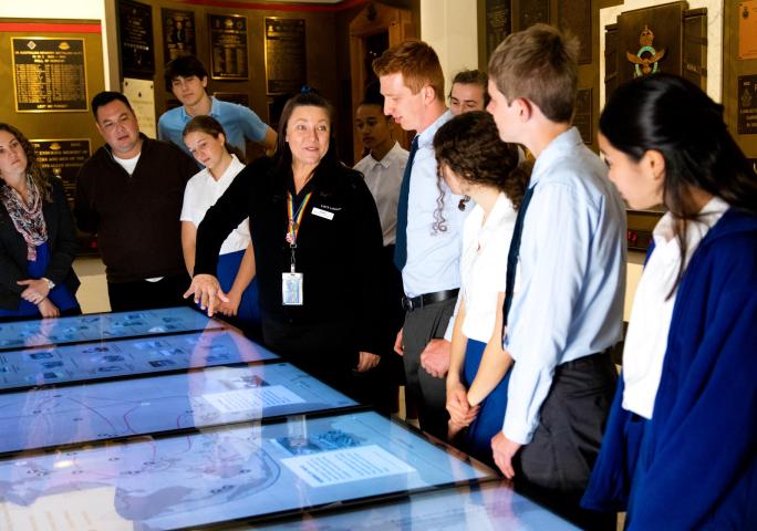 A group of students gathered around a staff member as she gestures to a touch screen in the Memorial Galleries. 