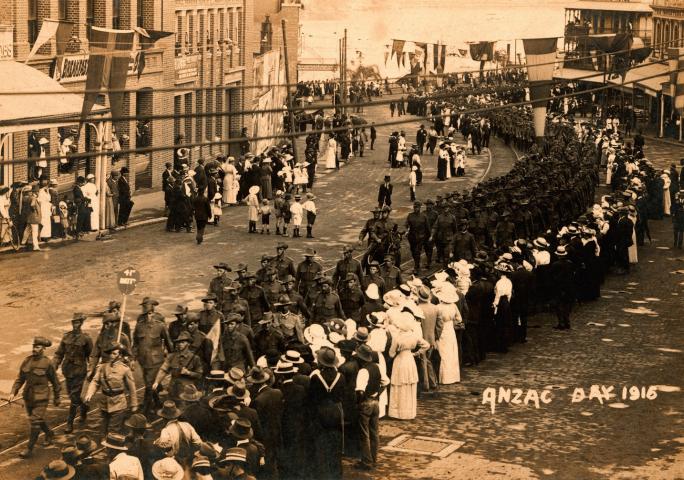 Old photograph from above of people gathered on the side of a street watching a Battalion march.