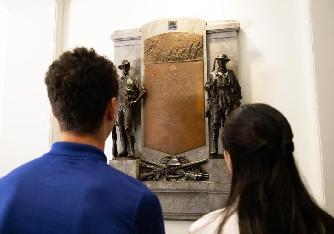 The back of two figures looking at a plaque featuring two soldiers in the gallery.