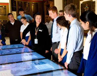 A group of students gathered around a staff member as she gestures to a touch screen in the Memorial Galleries. 